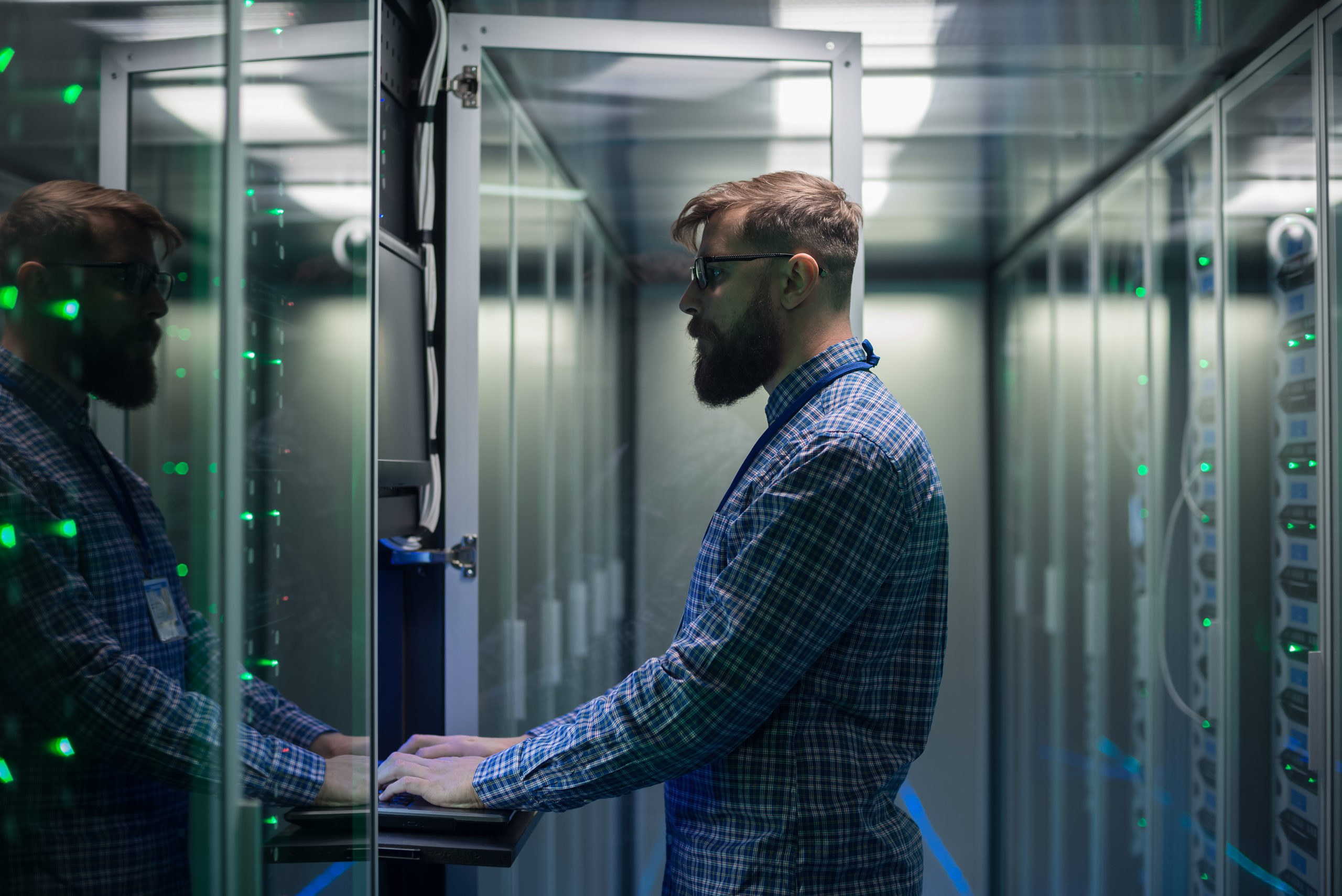 Adult bearded system administrator in glasses working on laptop and correcting errors in server hardware